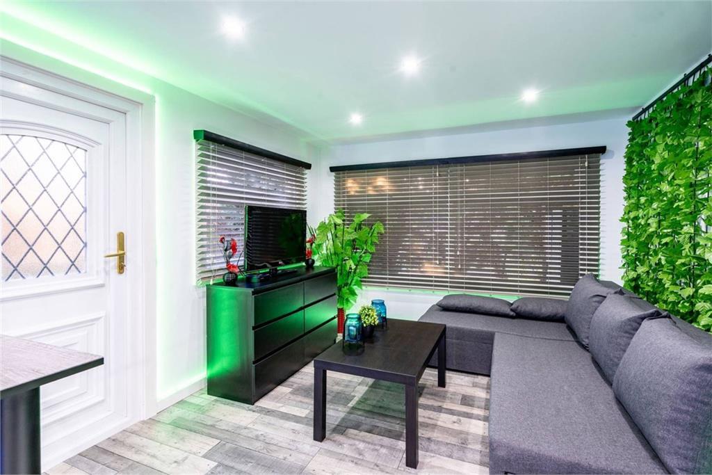 A small but modern living room with white walls, grey sofa, black tv unit, and green lighting.