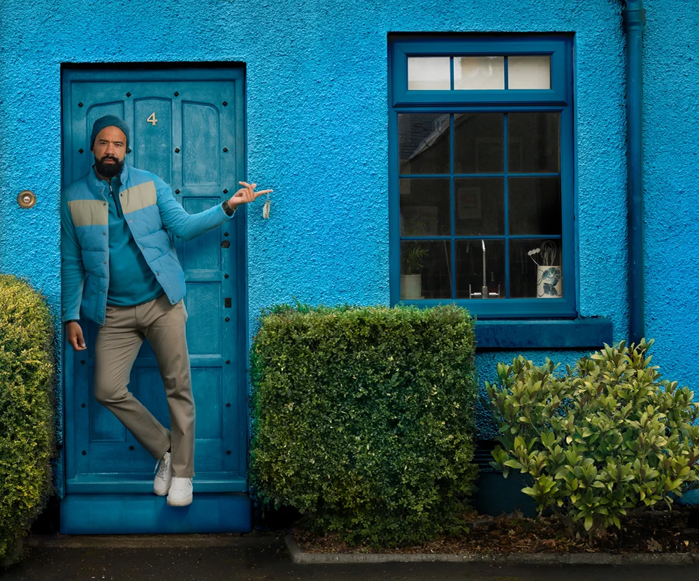 A man in a blue jacket leans against a blue door of a blue house, holding a key, with bushes on either side and a window to his left.