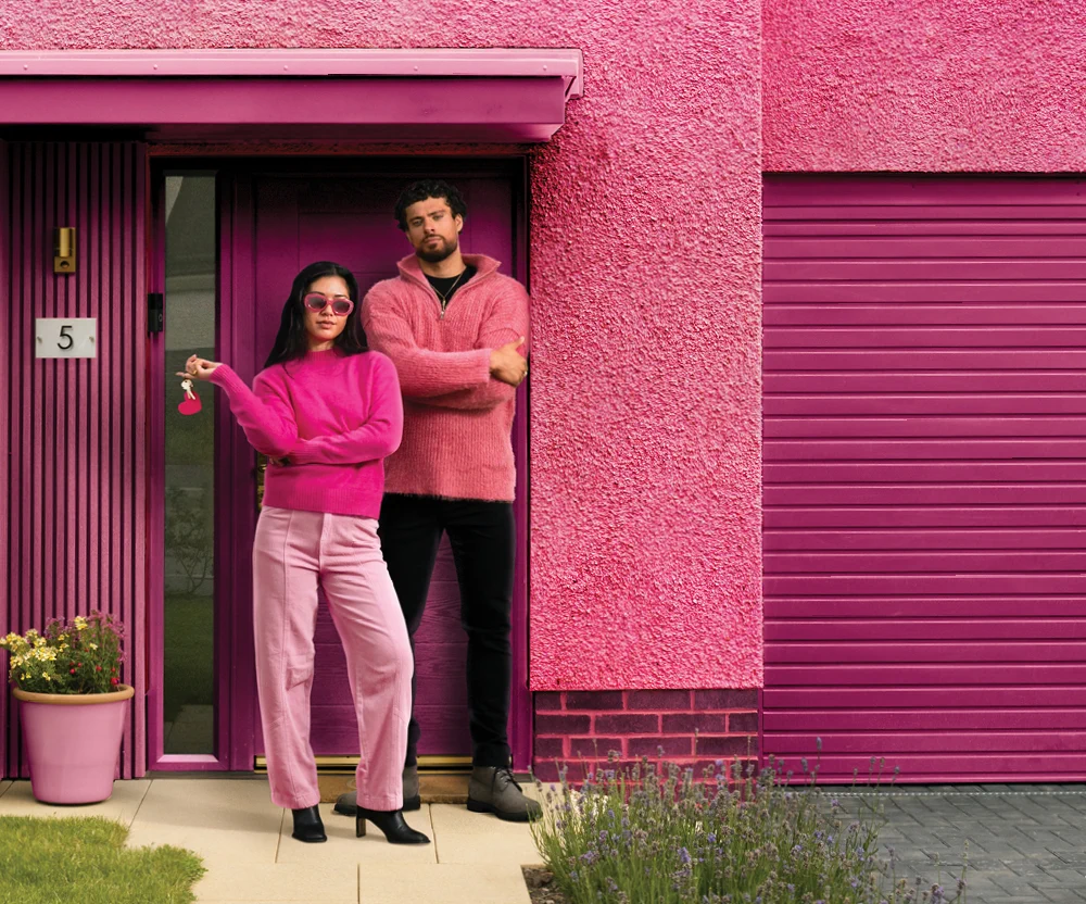 A woman holding house keys and a man stand in front of a bright pink house with a matching garage door.